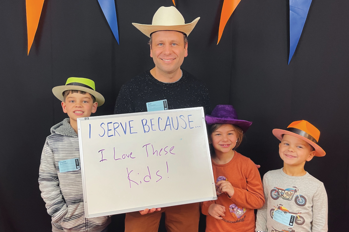 A volunteer and his family. They are wearing fun hats and holding a sign that says: I serve because I love these kids!